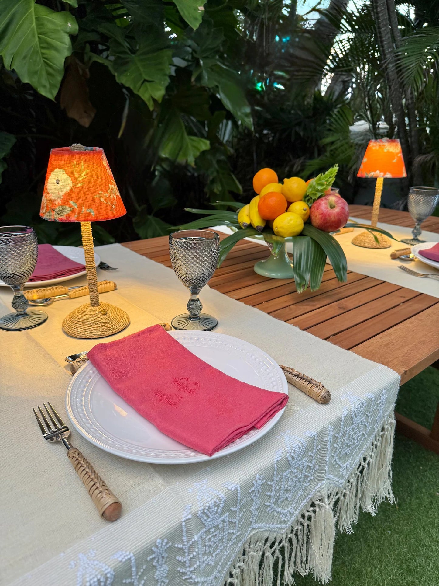 Outdoor dining setup featuring two glowing Tangerine floral lampshades on seagrass cordless lamps, surrounded by greenery and a vibrant fruit centerpiece.