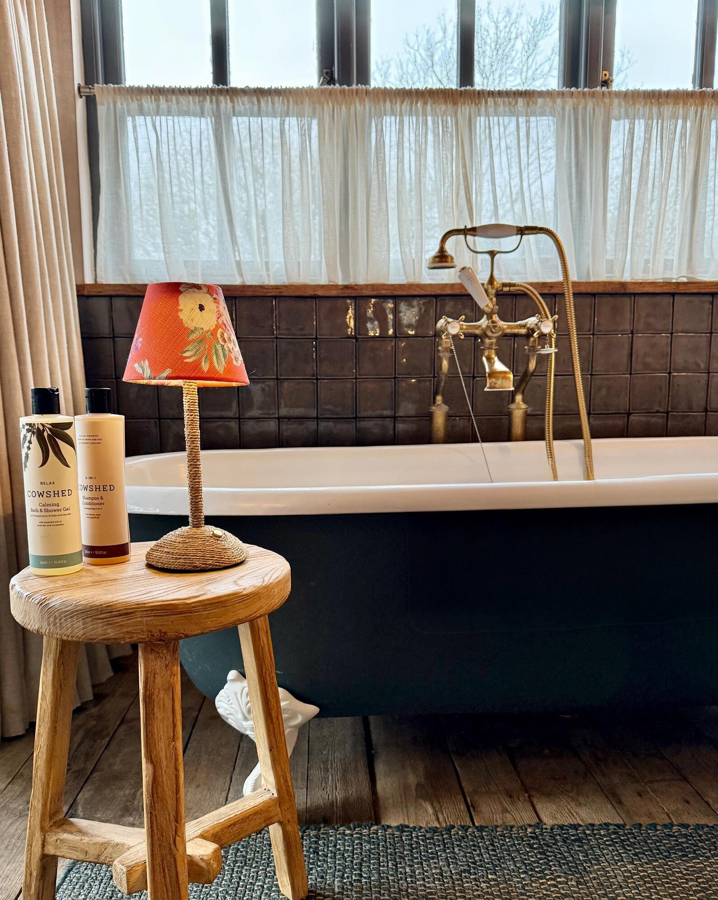 A coastal-style cordless lamp with a woven seagrass base and orange floral shade sitting on a wooden stool in a luxury bathroom next to a vintage bathtub.
