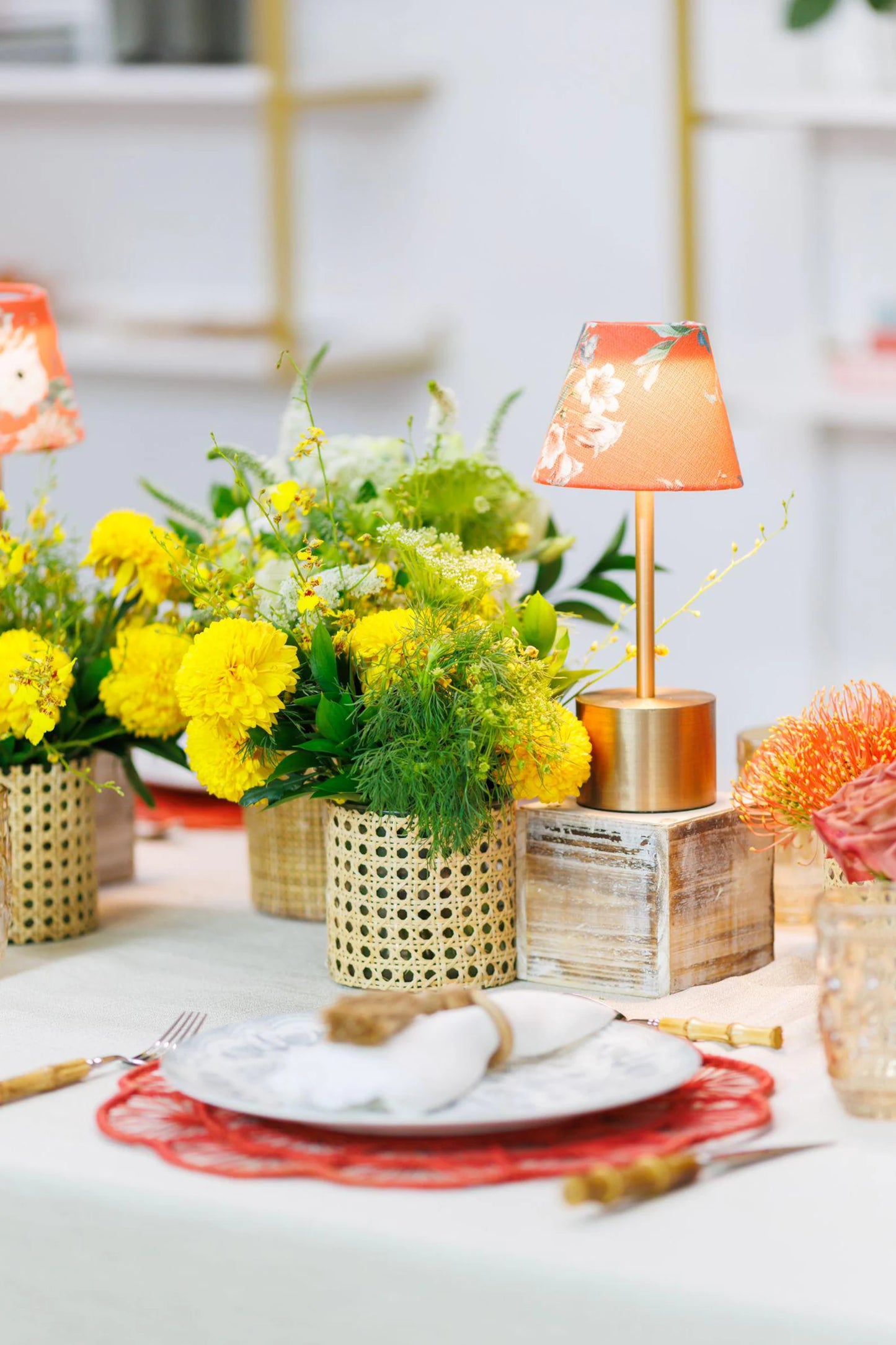 A gold-base cordless lamp with a floral orange shade styled on a wooden block amidst vibrant yellow flower arrangements and cane-textured vases