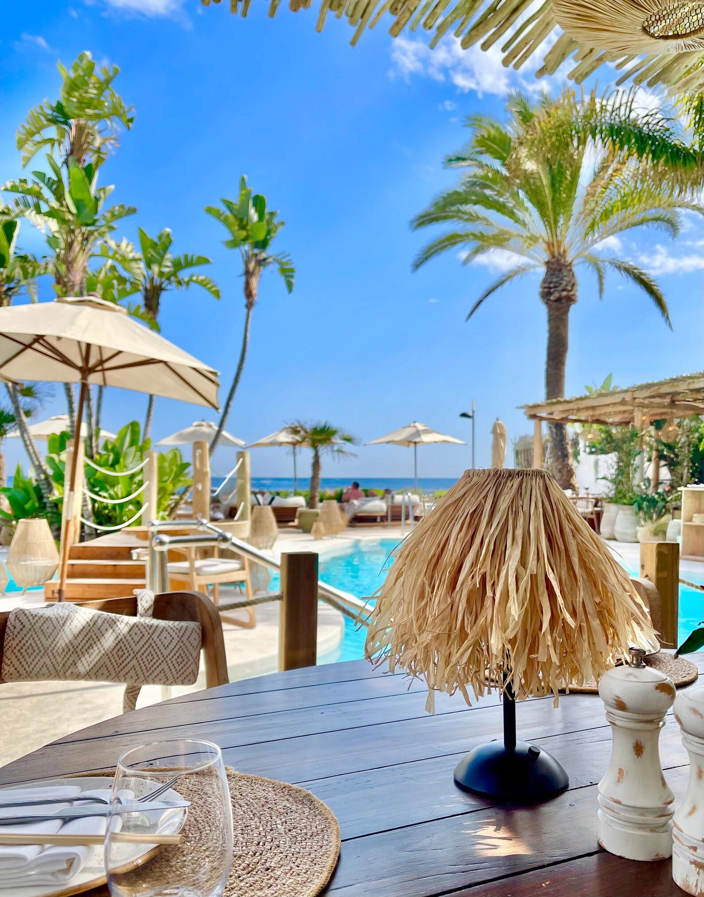 Breezy natural raffia lamp shade on a cordless base styled at a luxury beach club pool with palm trees and ocean views.