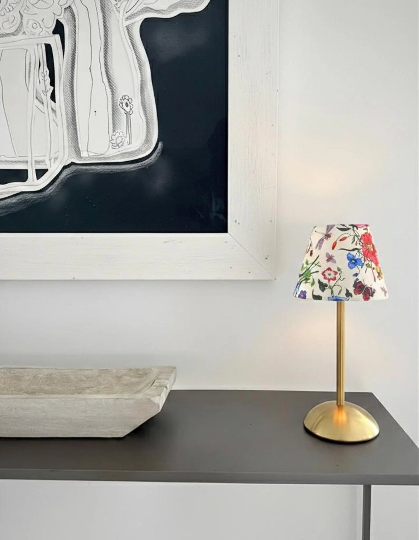 The Botánica botanical shade on a gold cordless lamp base styled on a minimalist black console table beneath a large art piece