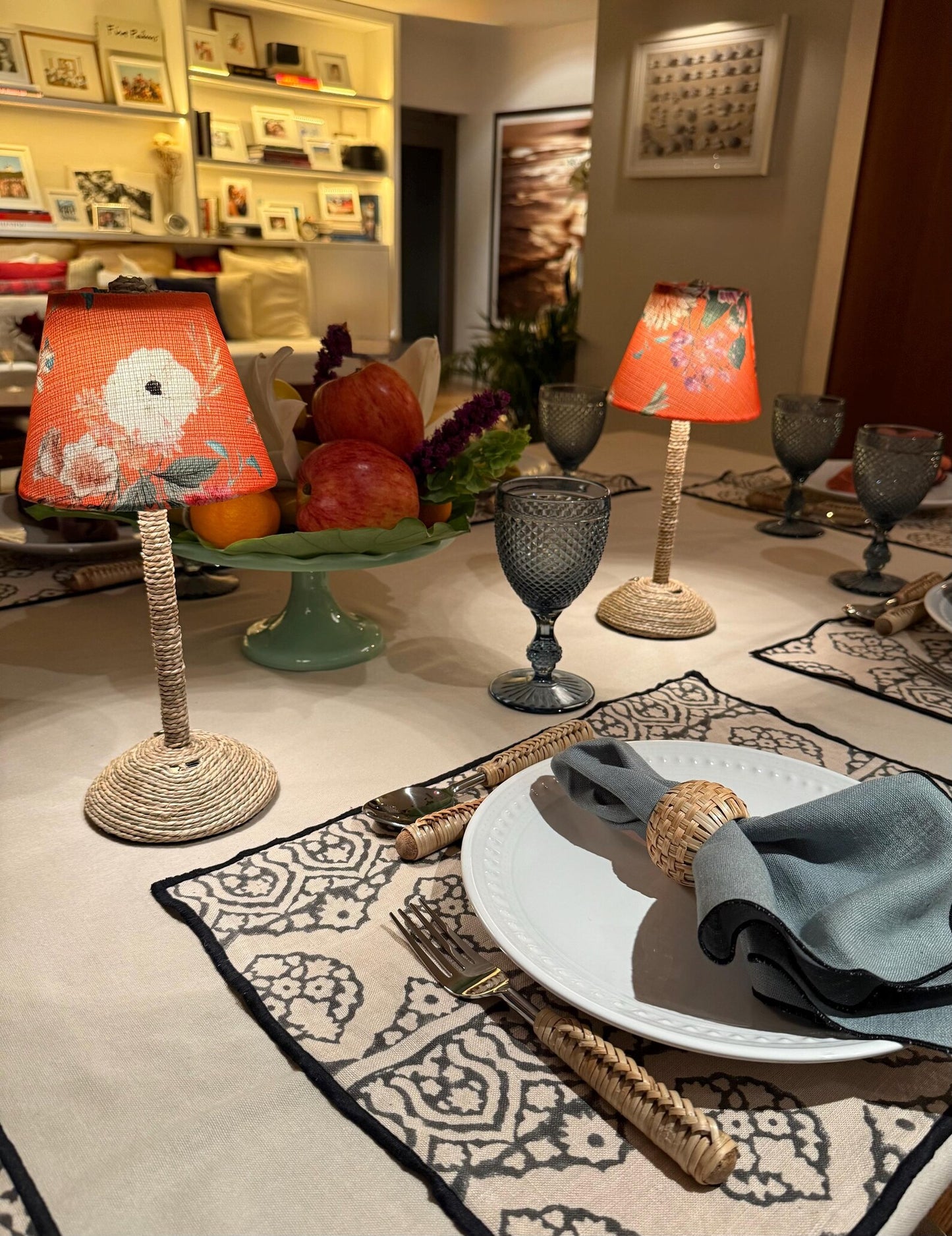 Elegant indoor dining table set with two glowing Tangerine floral lampshades on seagrass cordless lamp bases, surrounded by woven placemats, rattan-handled cutlery, and blue goblets, creating a cozy and inviting atmosphere