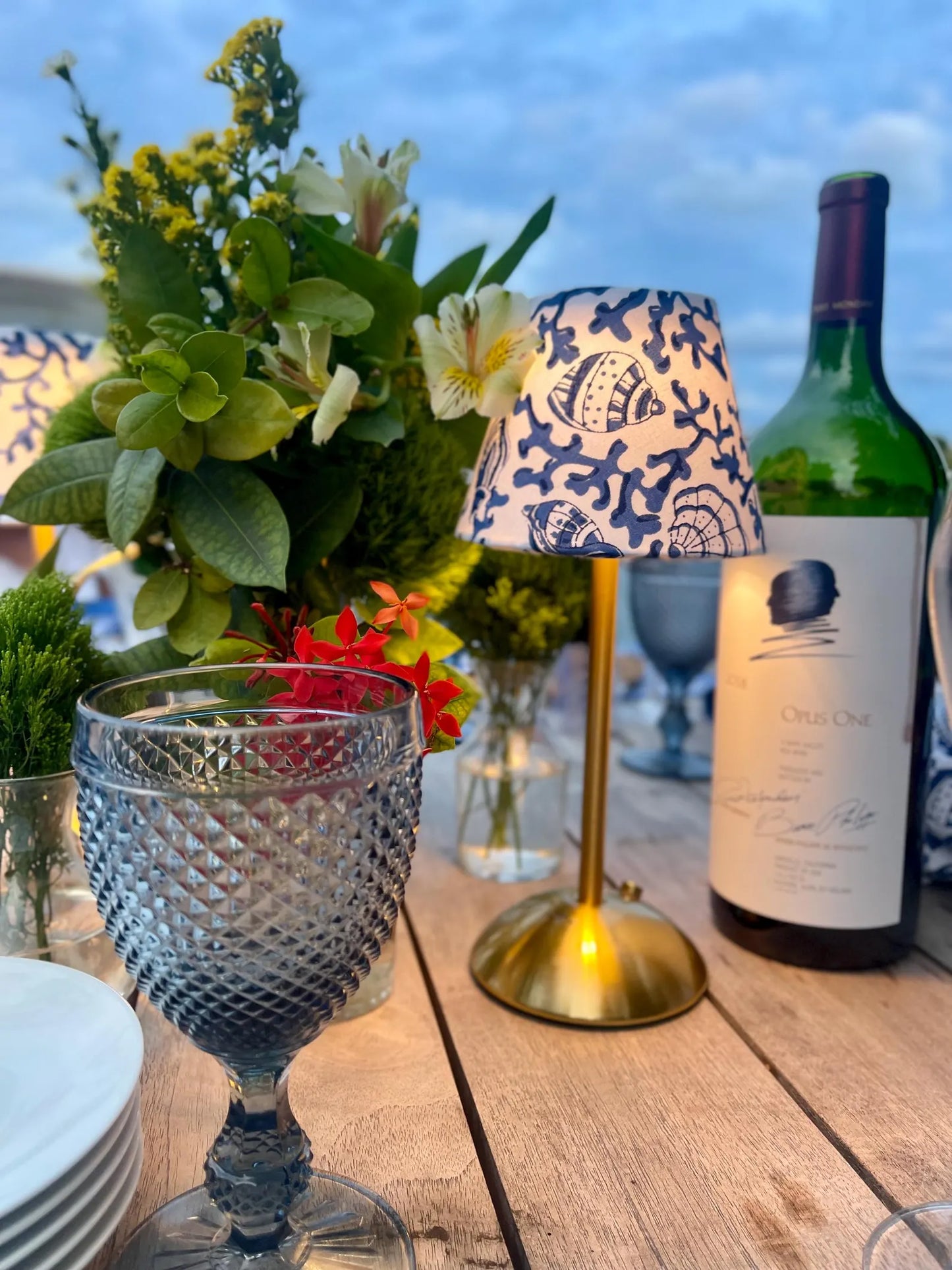 Handcrafted seashells print lampshade on a gold cordless lamp styled with an Opus One wine bottle for outdoor dining.