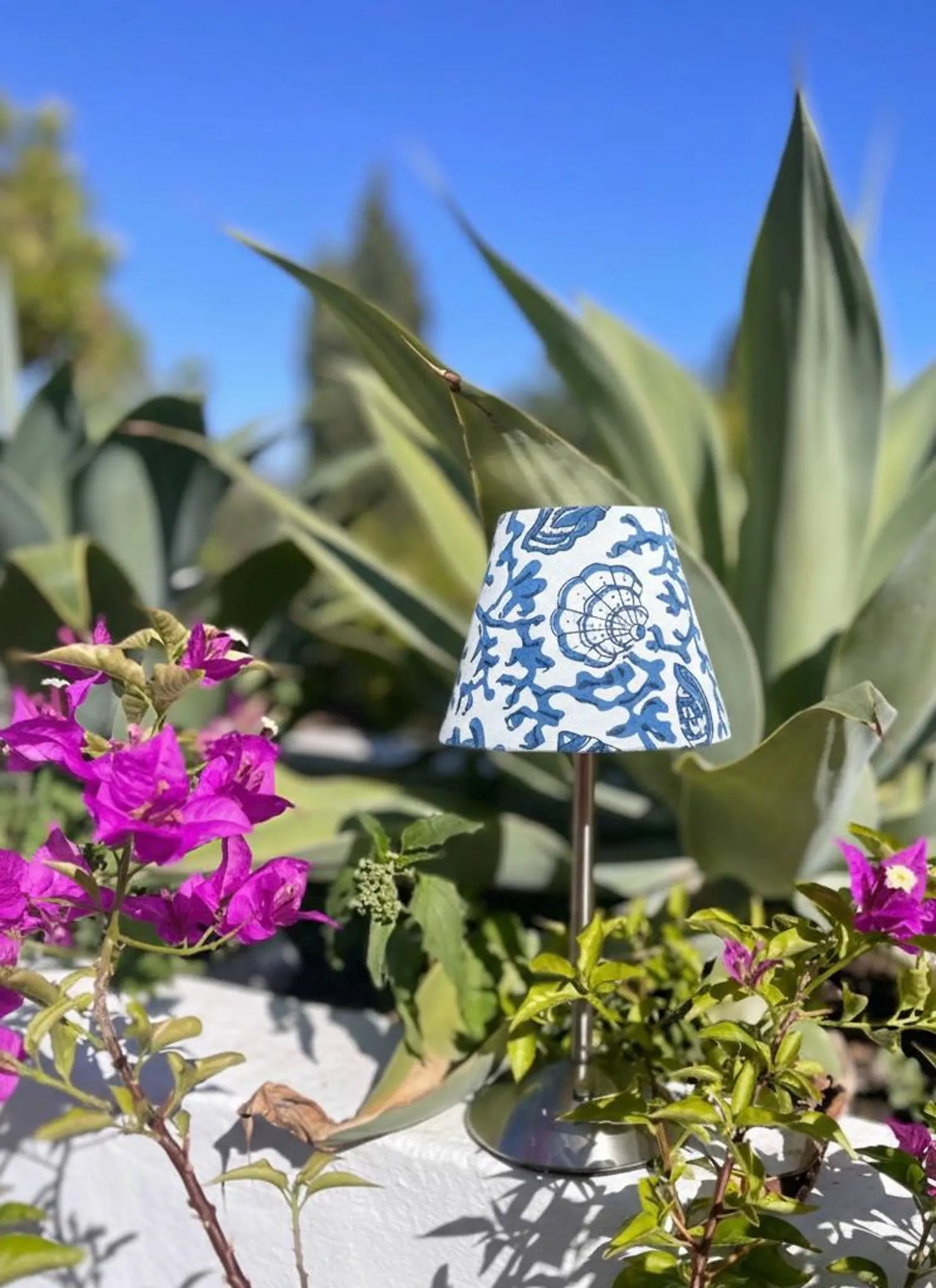 Blue seashells lamp shade on a silver cordless lamp styled outdoors with agaves and vibrant pink bougainvillea.