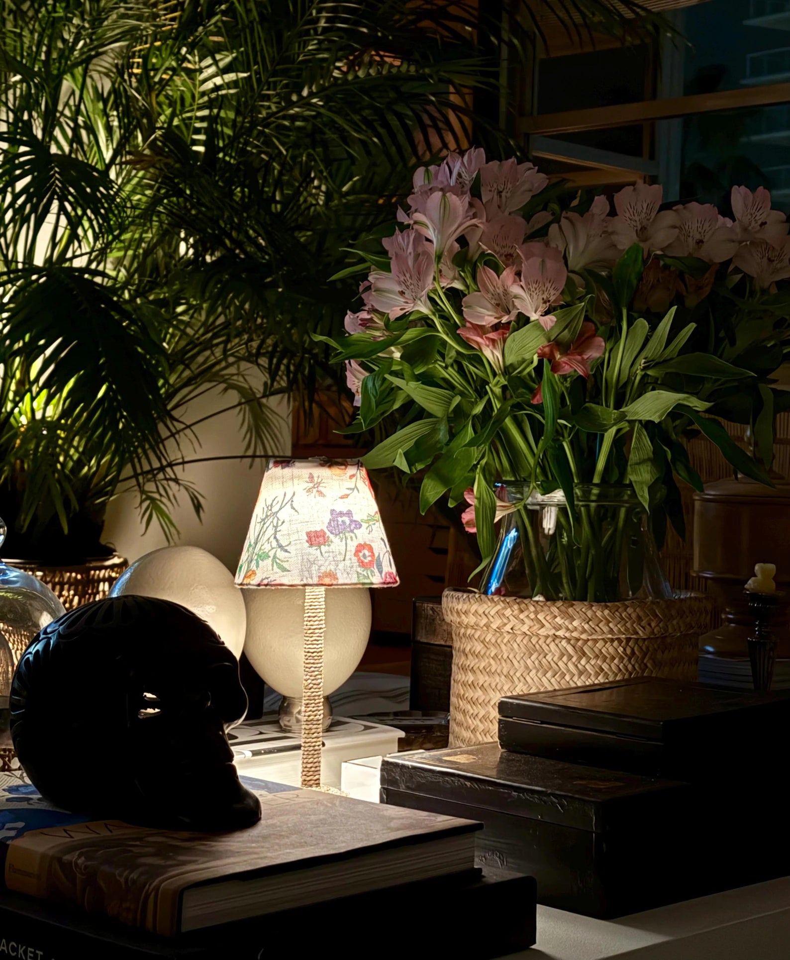 A low-light evening tablescape featuring The Botánica cordless lamp shade glowing softly next to pink lilies and a decorative black skull.