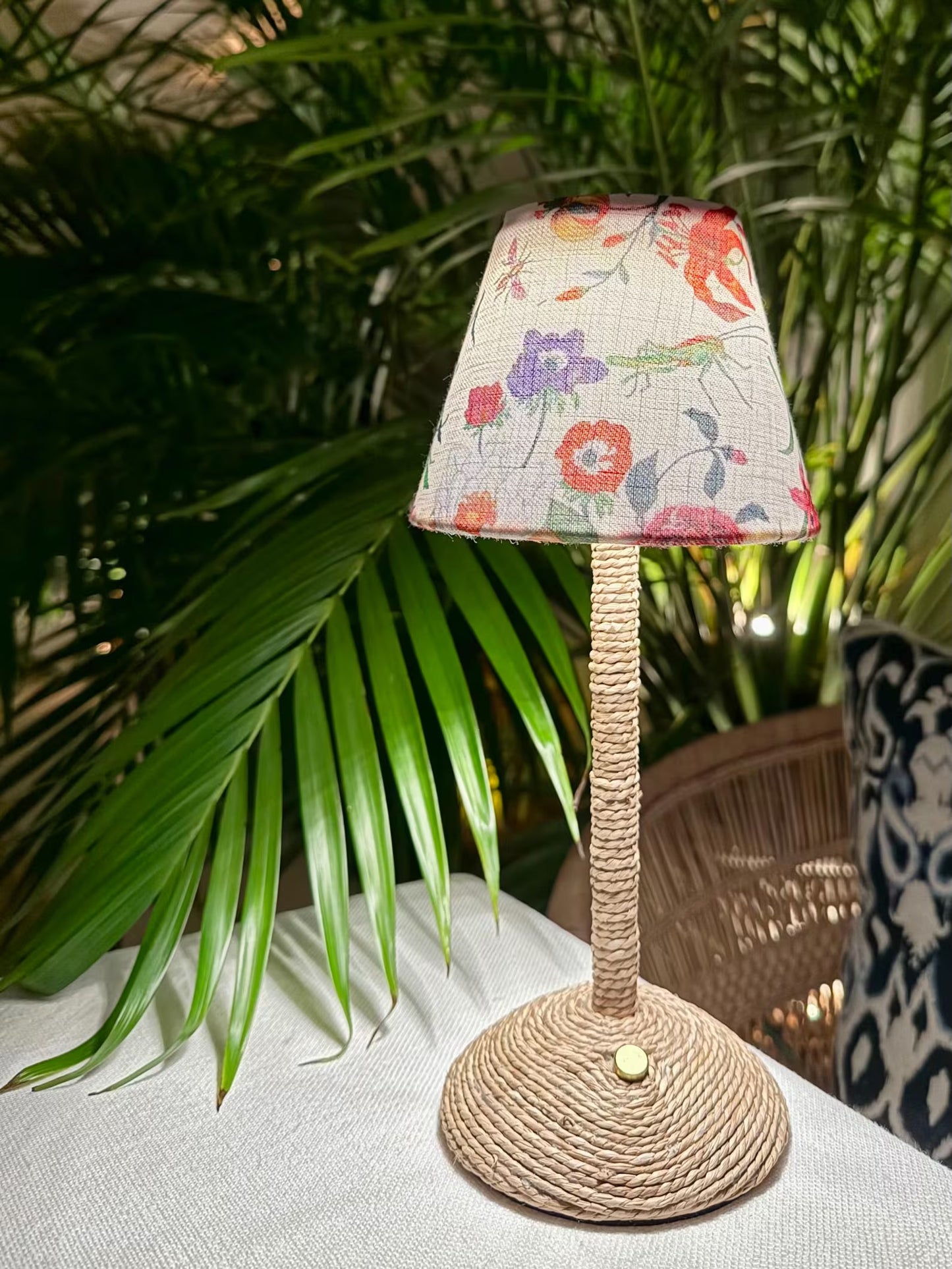 The Botánica floral shade on a seagrass cordless lamp base styled on an outdoor dining table with woven wicker chairs.