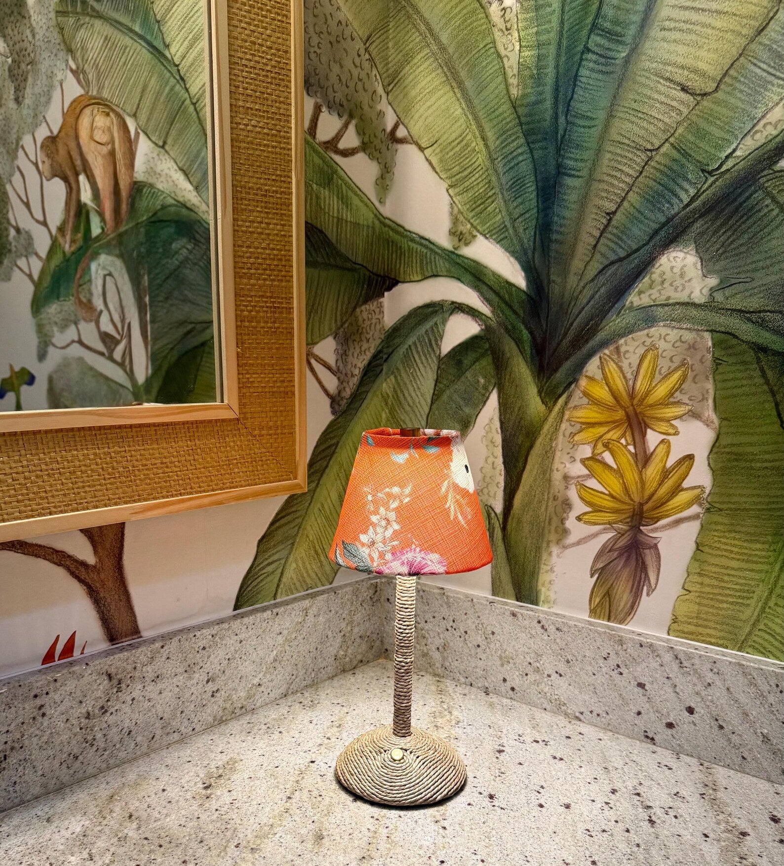 A cordless lamp with a seagrass base and orange floral lampshade placed on a marble countertop against a lush tropical monkey-themed wallpaper.