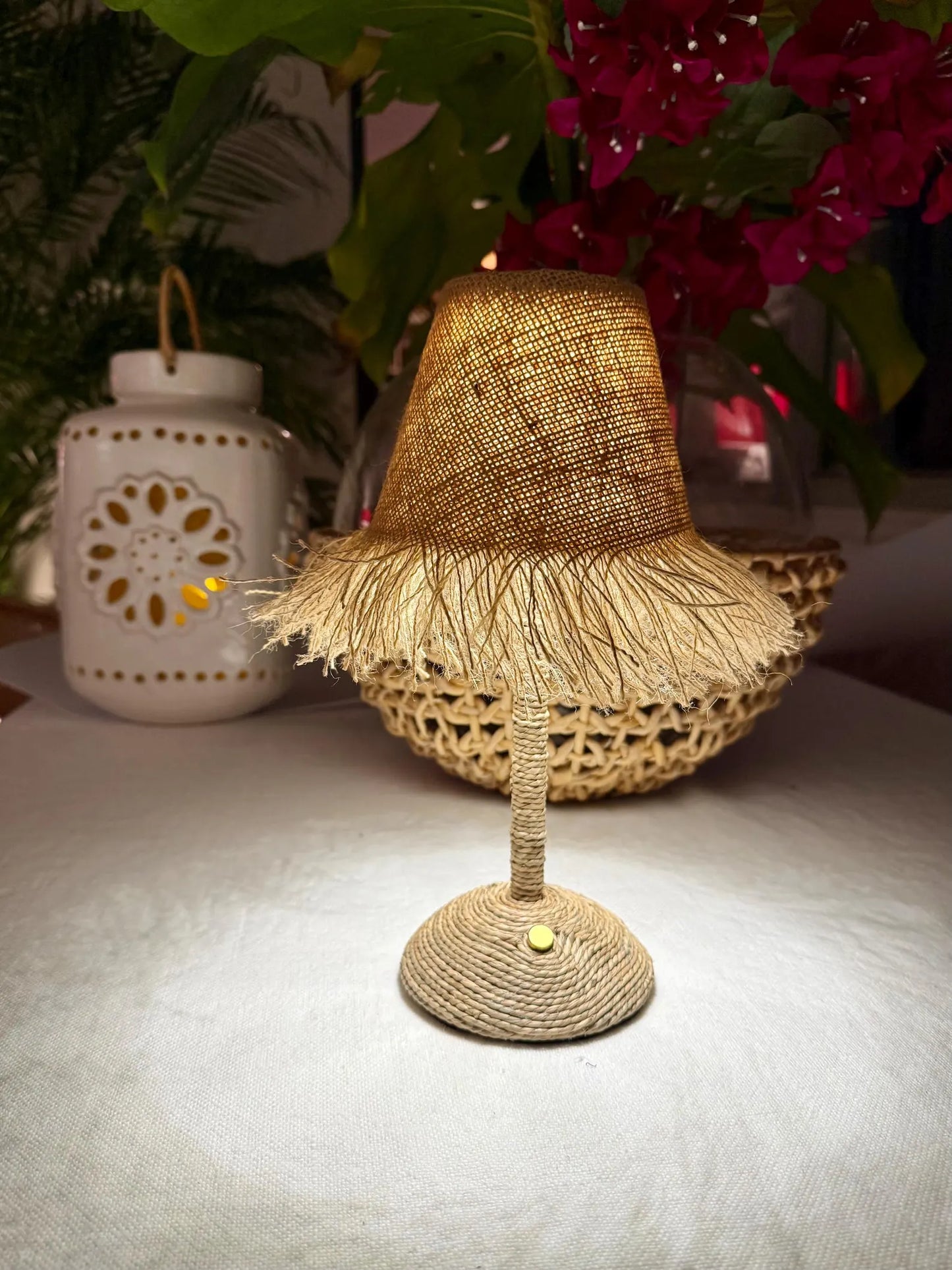 Lit burlap fringe lampshade on a woven cordless base styled with warm light and tropical pink flowers in a cozy interior setting.