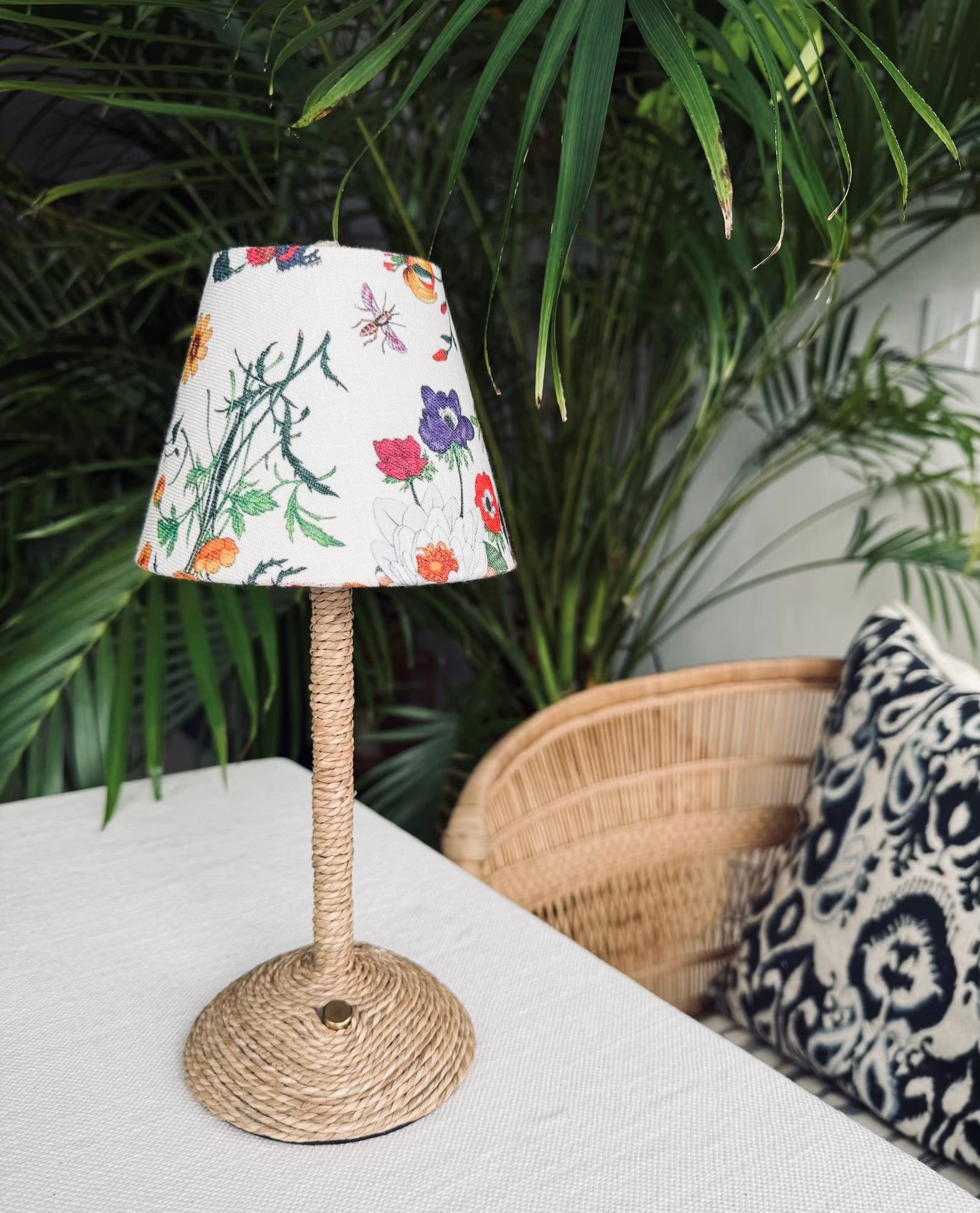 The Botánica botanical lamp shade styled on a seagrass-wrapped cordless lamp base with lush palm greenery in the background.