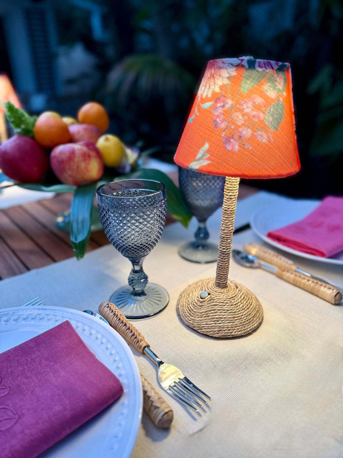 Close-up of a small cordless lamp with an orange floral lampshade and a seagrass-wrapped base on a dinner table next to a blue textured glass.